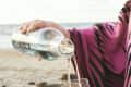 Person pouring water from a bottle into a glass on a sandy beach, wearing a pink and black patterned garment.