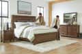 Two men making a bed in a modern bedroom with wooden furniture, striped bedding, and decorative vases on a dresser.