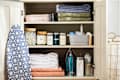 Linen closet with folded towels, labeled jars, cleaning supplies, and an ironing board with a blue geometric cover.