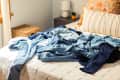 Pile of blue denim clothes on a bed with striped pillows and a wicker basket nearby.