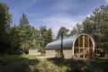 Curved-roof cabin with large windows surrounded by trees in a forest setting.
