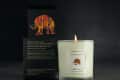 Lit gardenia tuberose candle in glass jar with colorful elephant design, next to matching black box on dark surface.