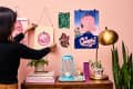 Woman hanging art on a pink wall with colorful prints, books, plants, a blue lantern, and a gold lamp on a wooden table.