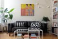 Black leather sofa with geometric pillows, large plants, modern art, and bookshelves in a bright living room.