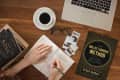Hands writing in a journal on a wooden desk with a coffee cup, glasses, photos, and "The Bullet Journal Method" book.
