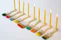 Paintbrushes with colorful bristles holding lit yellow candles in a row.
