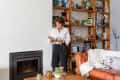 Woman reading a book by a fireplace, with a leather sofa and wooden bookshelves filled with decor and books in the background.