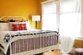 Bright bedroom with yellow walls, white metal bed, red patterned pillow, and a floor lamp by sheer curtained windows.