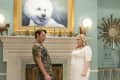 Two people stand in a room with a large dog portrait, ornate mirror, and decorative horse figurines on the mantel.