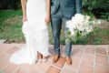 Bride in lace gown holding dress, groom in gray suit with brown shoes, holding white floral bouquet on terracotta tiles.