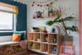 Cozy kids' room with wooden shelves, colorful toys, a large plant, and a rainbow cushion on a polka dot wall.