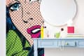 Pop art mural of a woman's face next to a blue desk with a round mirror and yellow pendant lights.