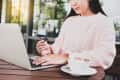 Woman in pink sweater holding credit card, using laptop at a wooden table with a cup of coffee.