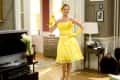 Woman in a bright yellow dress and gloves standing in a living room with a TV, flowers, and a lamp.