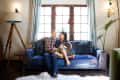 Couple sitting on a blue patterned sofa in a cozy living room with large windows, a tripod lamp, and a potted plant.