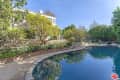 Curved swimming pool surrounded by stone path, lush greenery, and a two-story house.