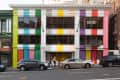 Colorful building facade with vertical stripes in green, blue, yellow, orange, purple, and red, with cars parked in front.