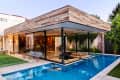 Modern glass-walled house with wooden accents, pool, and patio chairs on a lawn.