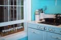Retro kitchen with blue cabinets, white stove, hanging light, spice rack by window, and colorful teapots on a shelf.