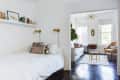 A small bedroom that has a floating shelf stacked with books is connected to a living room