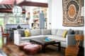 Living room with gray sectional sofa, colorful cushions, wooden coffee table, leather poufs, and decorative wall tapestry.