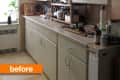 Cluttered kitchen with beige cabinets, countertop appliances, and a window with a potted plant.
