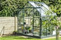 Glass greenhouse in a garden with potted plants and wooden fence, surrounded by lush greenery.