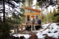 Couple standing on porch of a small wooden cabin in snowy forest, surrounded by pine trees.