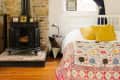 Cozy bedroom with a wood stove, brick wall, colorful bedspread, yellow pillows, and dark curtains.