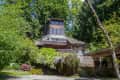 Wooden house with large windows surrounded by lush greenery and trees, featuring a covered patio area.