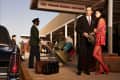 Man in suit and woman in colorful dress at TWA airport terminal, with luggage and a porter nearby.