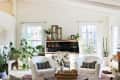 Bright living room with white armchairs, black patterned cushions, wooden desk, and various potted plants by large windows.