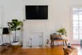 Living room with a white fireplace, wall-mounted TV, potted plants, and a wooden side table with books and decor.