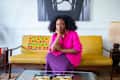 Woman in a bright pink blouse sitting on a yellow sofa with a colorful patterned throw, next to a lamp and magazines on a table.