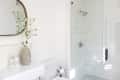 Bathroom with glass shower, white tiles, oval mirror, and vase with white flowers on the counter.