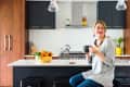 Woman sitting on a kitchen stool, holding a mug, with a fruit bowl on the counter and pendant lights above.