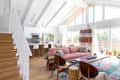 Open-concept living room with pink sofa, wooden chairs, colorful rug, and large windows overlooking greenery.