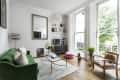 Living room with green sofa, glass coffee table, large windows, and wooden floor. Decor includes plants and a pink pouf.
