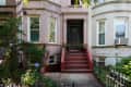 Brownstone entrance with red steps, black door, and potted plants, flanked by windows and greenery.