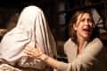 Woman in a beige sweater looking scared, touching a figure covered with a white sheet in a dimly lit room.