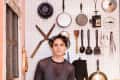 Person in a kitchen with pegboard wall displaying pots, pans, utensils, and a vintage rifle.