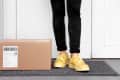Person in yellow sneakers standing on a doormat next to a cardboard package.