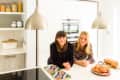 Two women smiling in a modern kitchen with pendant lights, bread, and croissants on the counter.