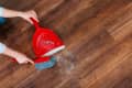 Hands sweeping dust on wooden floor with red dustpan and brush.