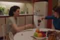 Woman in blue pouring drink for another woman at a kitchen table with fruit bowl and peanut butter jar.