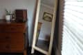 Wooden floor mirror leaning against a wall beside a wooden dresser with a potted plant and small chest.