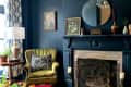 Dark blue living room with green armchair, patterned pillow, ornate fireplace, round mirror, and framed art.