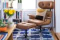 Brown leather lounge chair with ottoman on blue geometric rug, surrounded by bookshelves, yellow flowers, and a cactus.