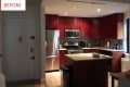 Dimly lit kitchen with red cabinets, stainless steel appliances, and a small island with a bowl and papers.