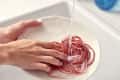 Hands washing a white plate with red silicone bands under running water in a sink.